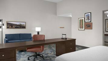 A hotel room with a bed, a desk, a chair, a sofa, a lamp, and framed pictures on the wall.