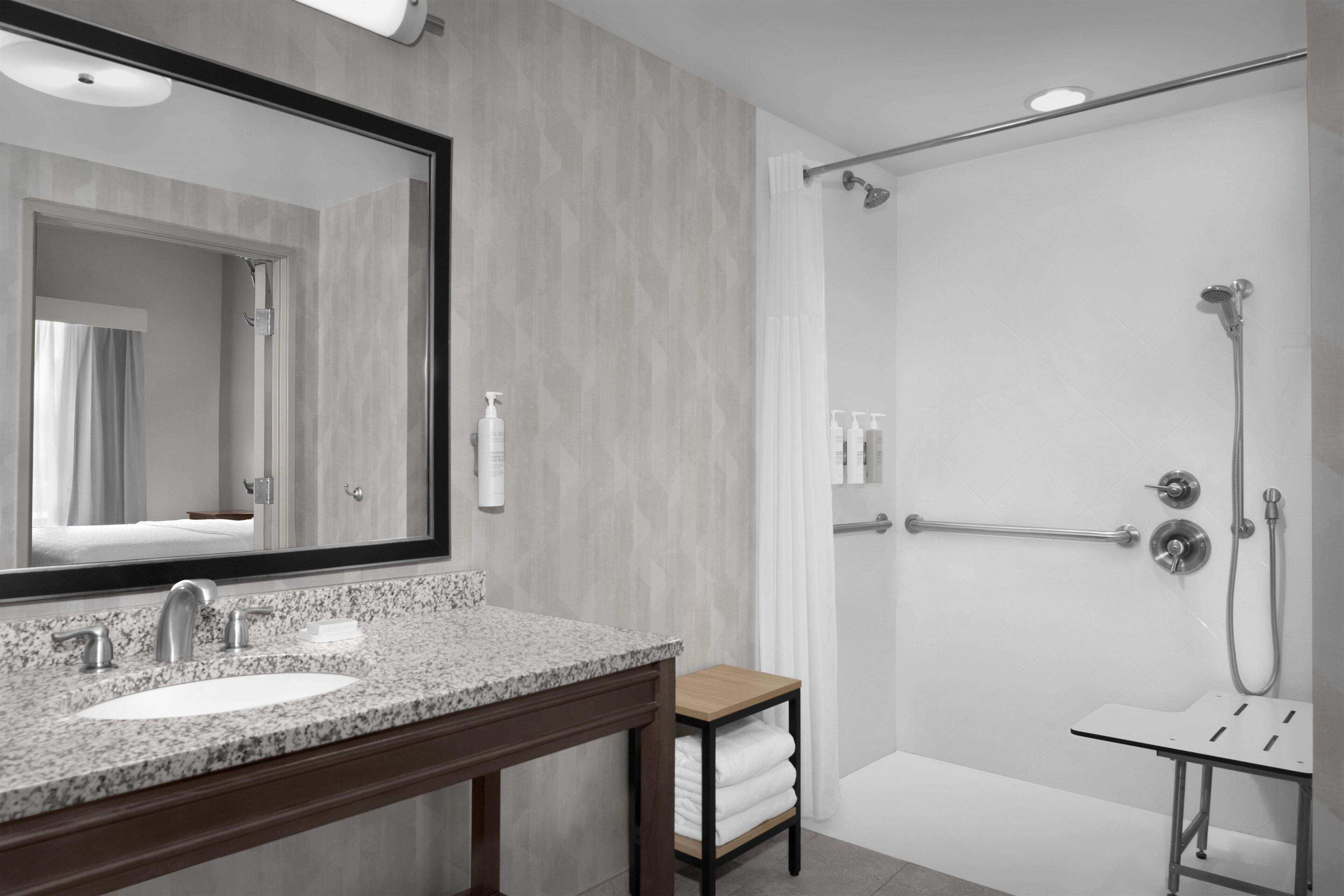 Une salle de bain moderne avec un lavabo surmonté d’un plan de travail en granit, un grand miroir, une douche équipée d’un pommeau de douche à main et un porte-serviettes.