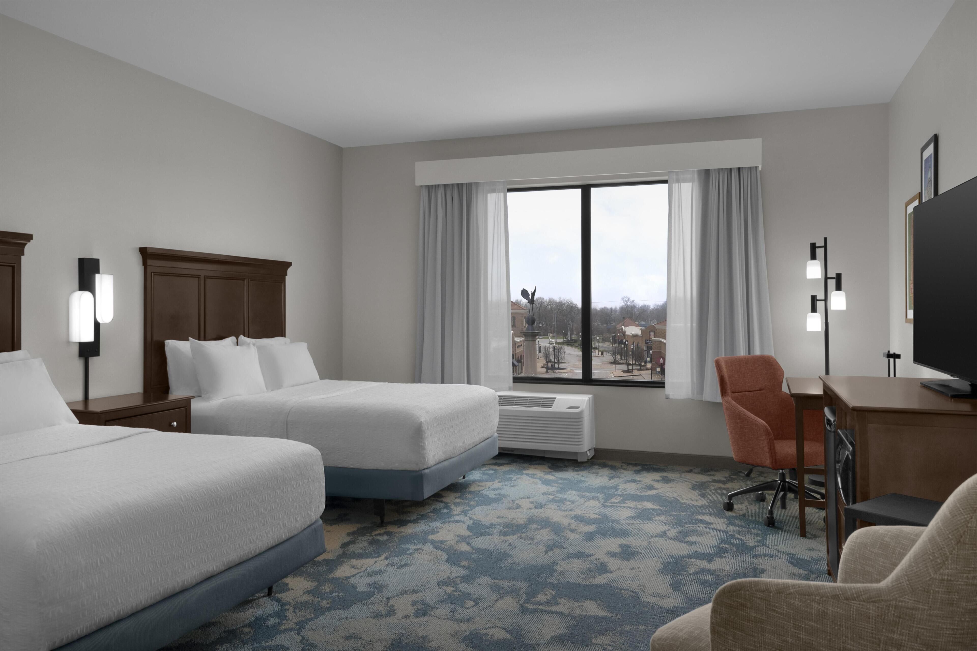 Ein Hotelzimmer mit zwei Betten, einem Schreibtisch, einem Sessel, einem Fernseher und einem großen Fenster mit Vorhängen.