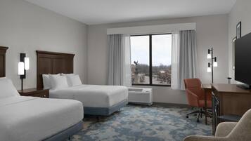 Ein Hotelzimmer mit zwei Betten, einem Schreibtisch, einem Sessel, einem Fernseher und einem großen Fenster mit Vorhängen.