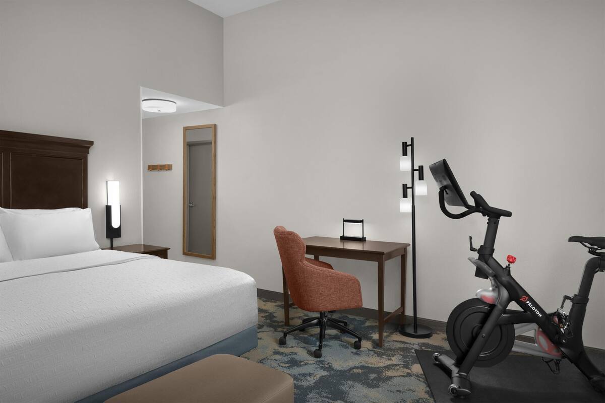 A hotel room with a bed, a desk, an office chair, a standing lamp, and an exercise bike.
