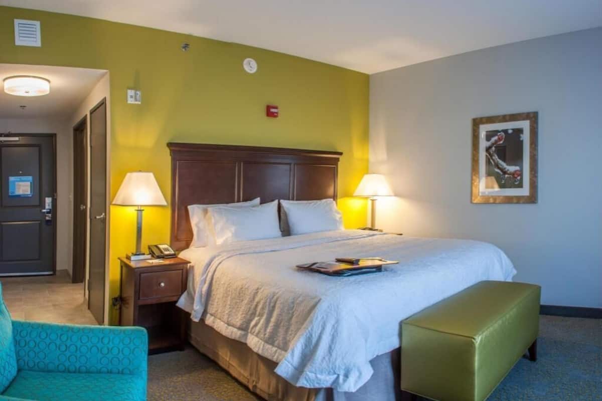 A hotel room with a bed, two bedside lamps, a nightstand, a green armchair, and a bench.