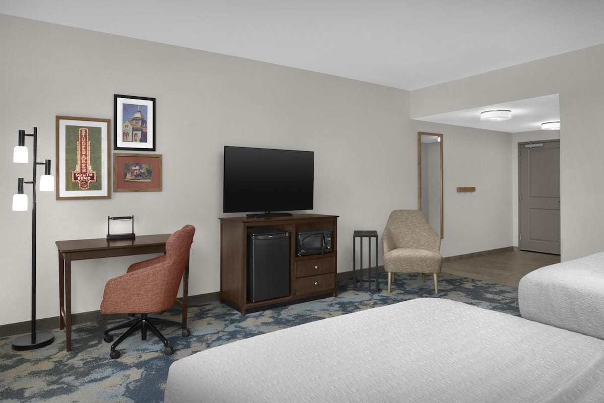 A hotel room with a bed, a desk with a television, a chair, and framed pictures on the wall.
