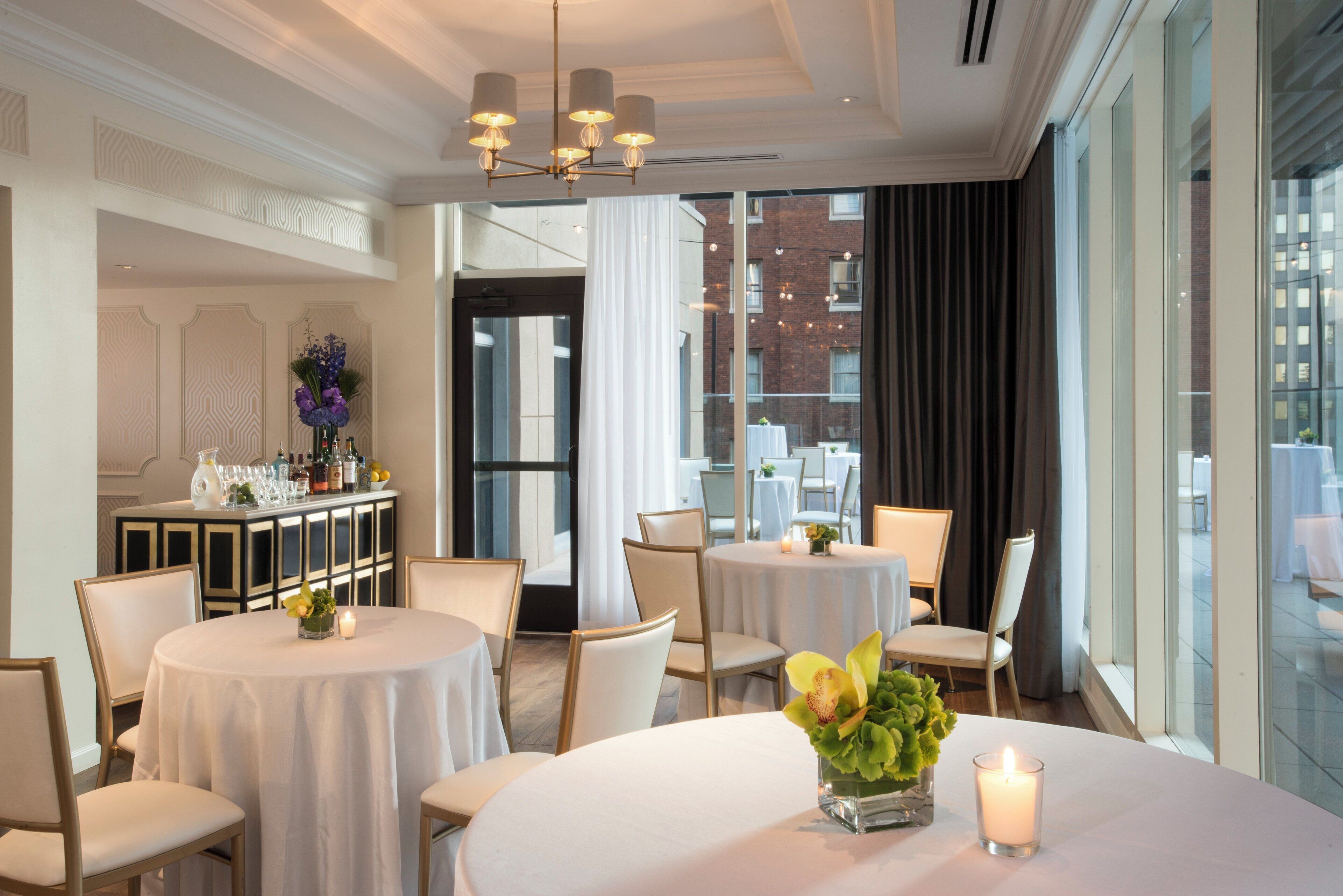 Une salle à manger d’hôtel avec des tables rondes, des chaises, un bar avec des bouteilles et une vue sur la ville grâce à de grandes fenêtres.