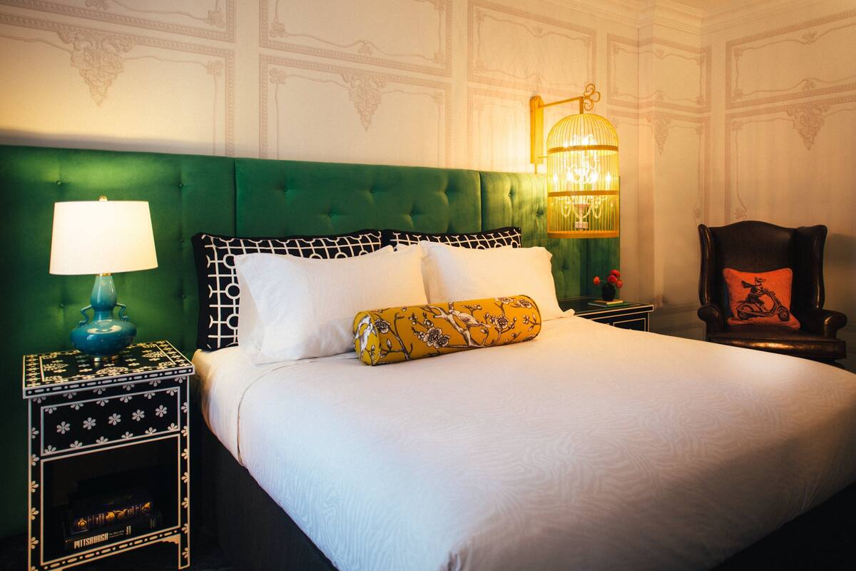 A neatly made bed with white linens and decorative pillows, a green upholstered headboard, a bedside table with a lamp, and a wall-mounted light fixture.