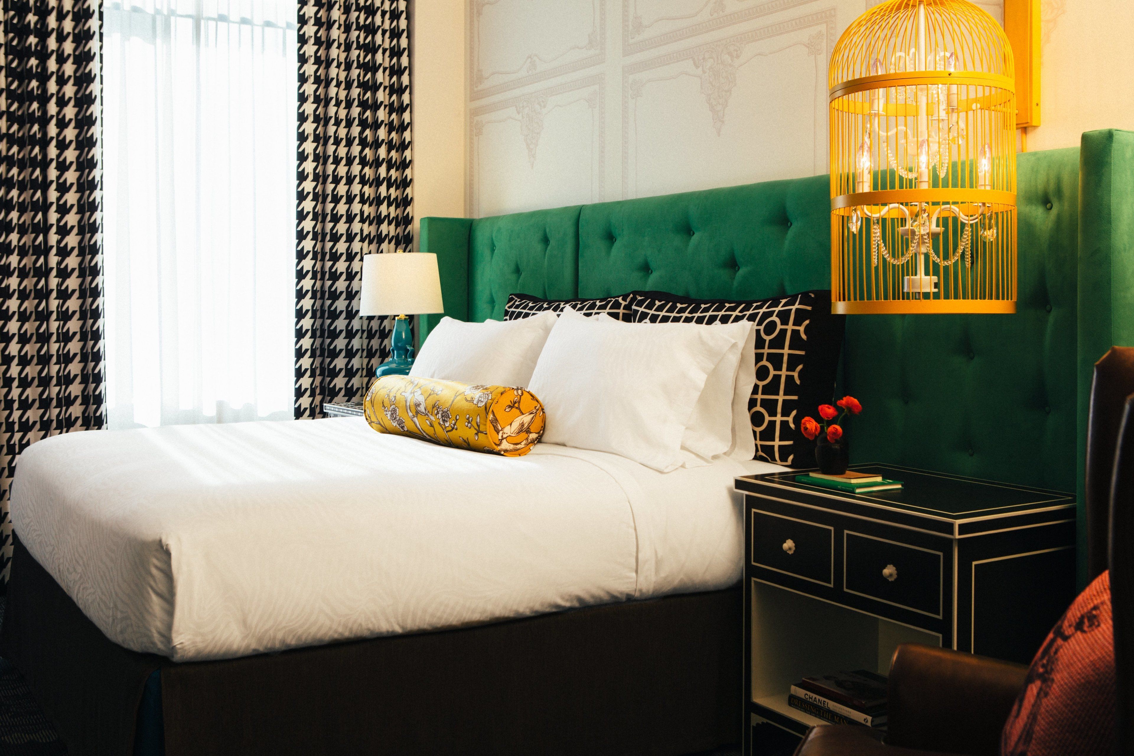A hotel room with a large bed, green upholstered headboard, a nightstand with a lamp, and a decorative birdcage.