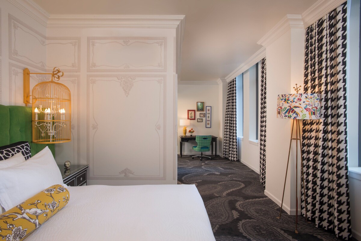 A hotel room with a bed, a green chair, a desk with a lamp, and a houndstooth curtain.