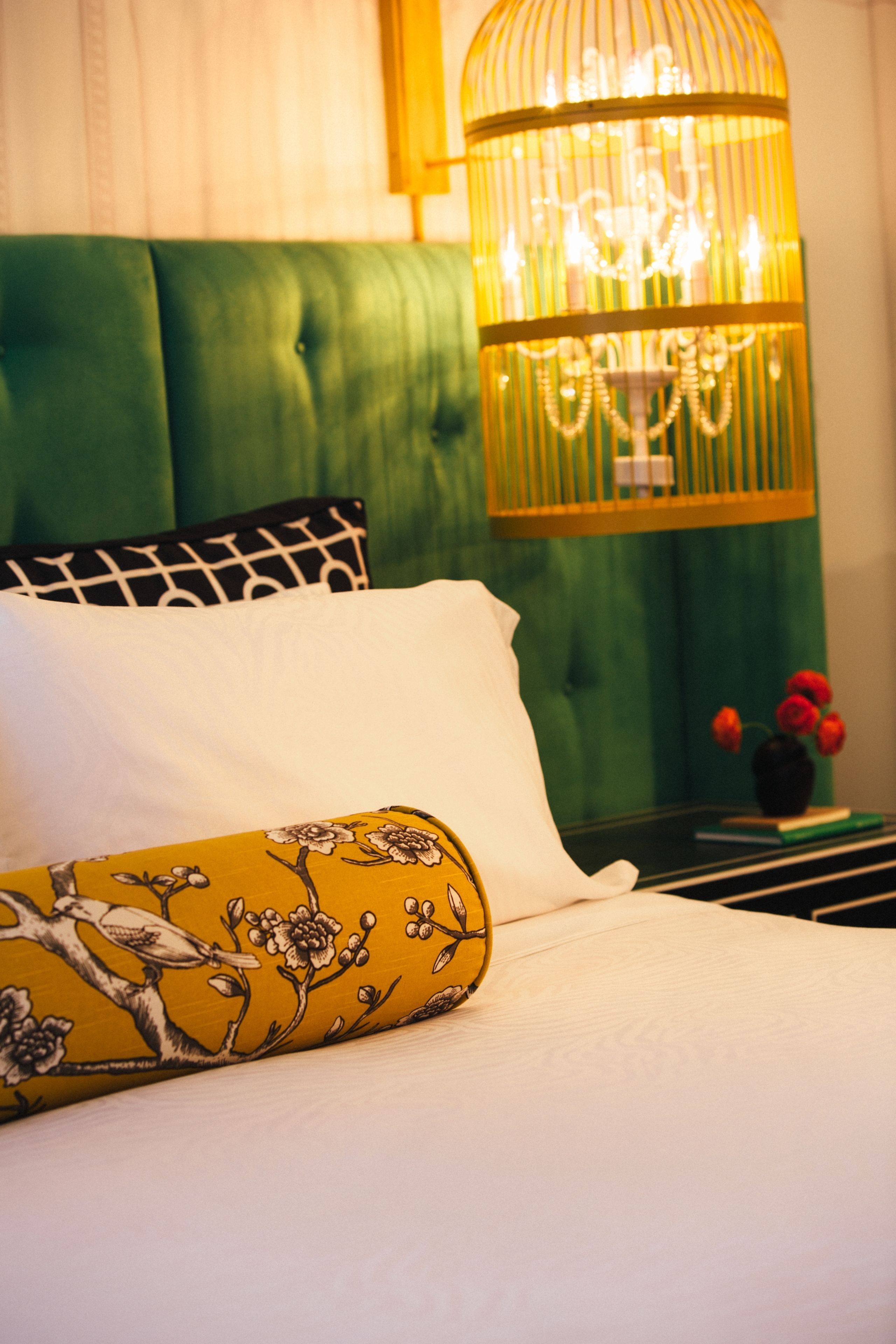 Cama com cabeceira verde, travesseiro amarelo com padrão de pássaros e flores, e um travesseiro branco. Cortina verde ao fundo e uma pequena mesa com um vaso de flores vermelhas.