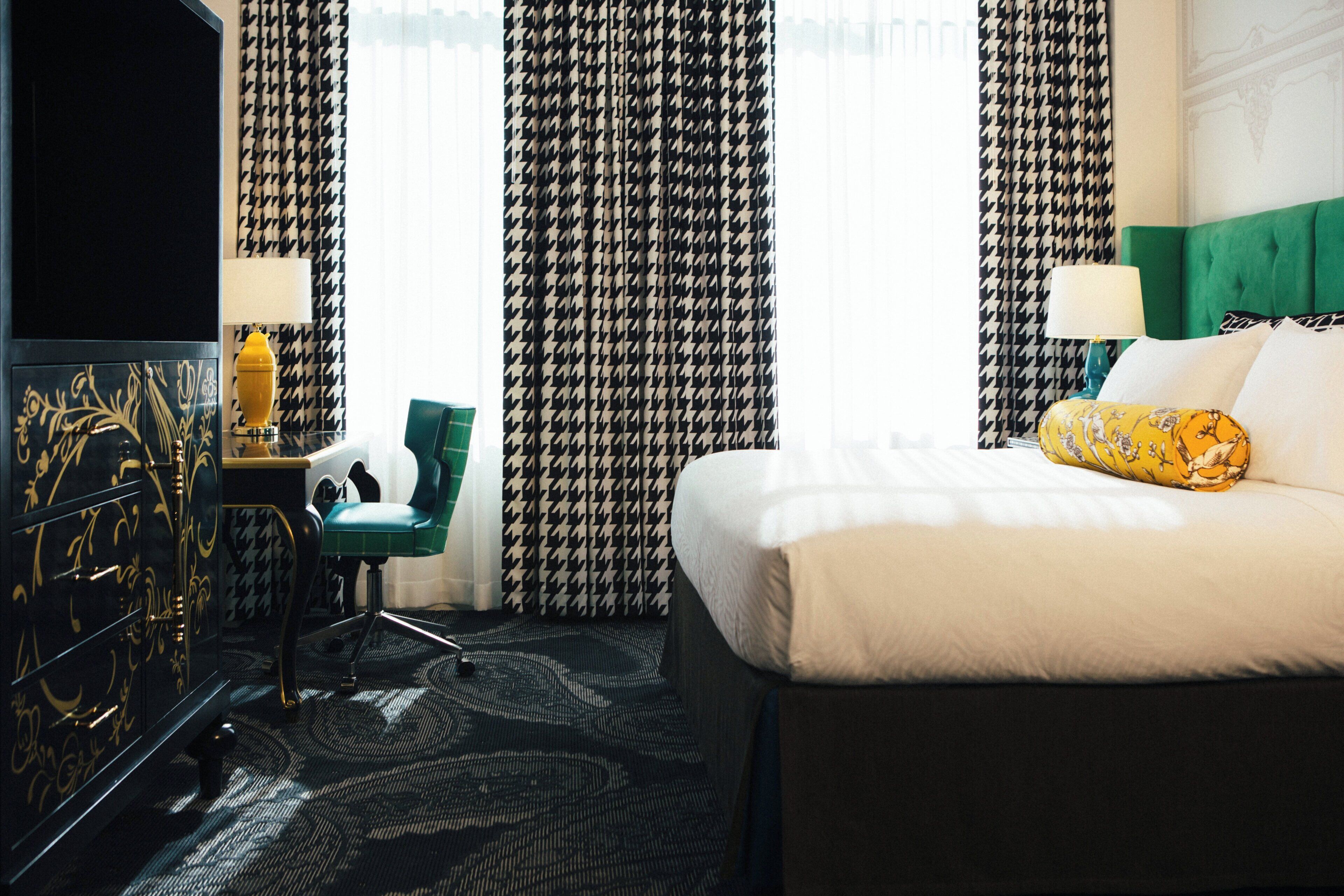 A hotel room with a bed, a green chair, a dresser, and patterned curtains.