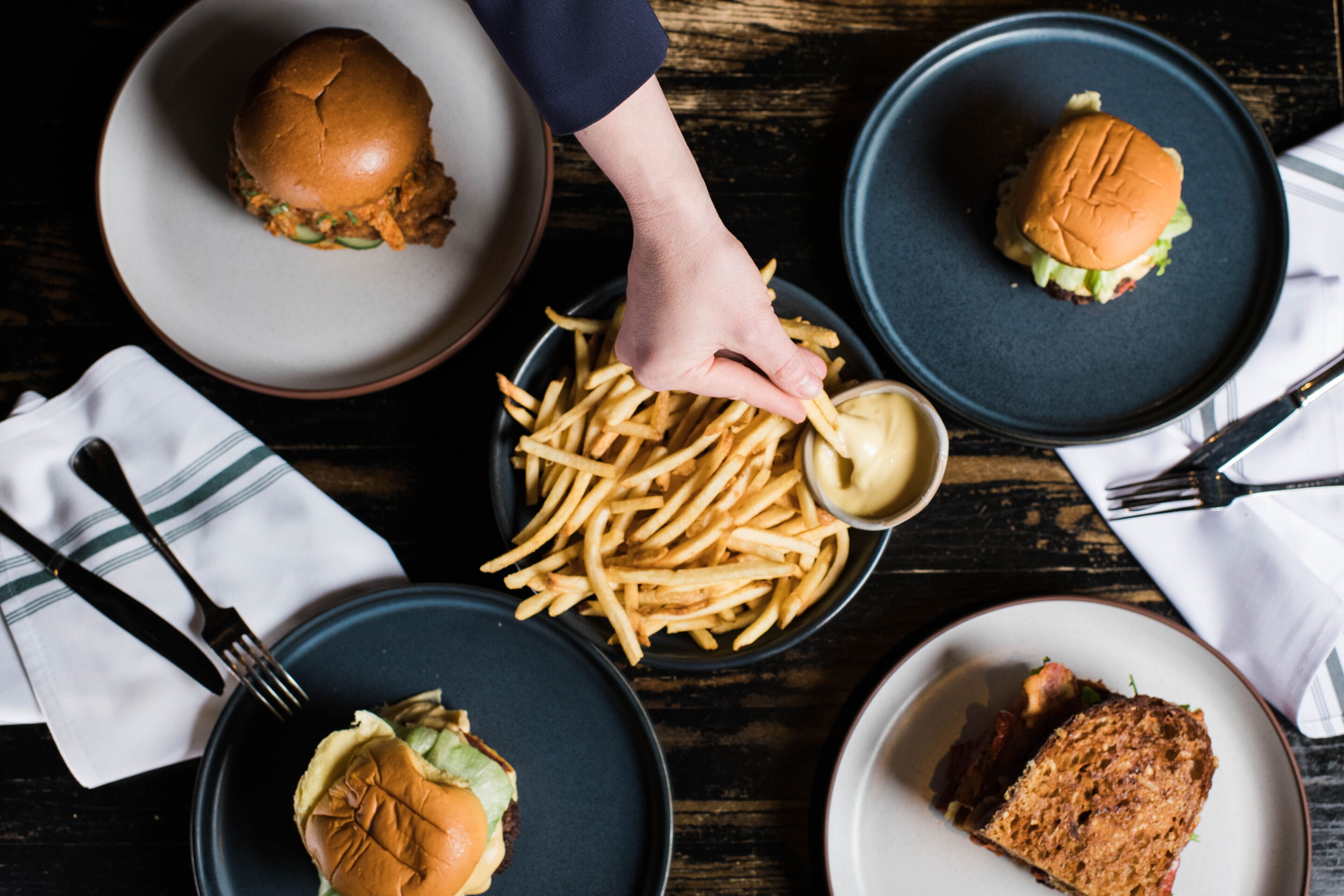 A dining table with four plates of food, including burgers and fries, and a hand reaching for the fries.