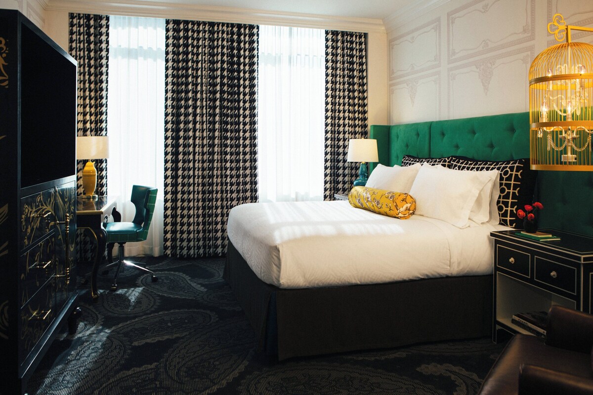 A hotel room with a large bed, a green headboard, a TV, a desk, and a lamp.