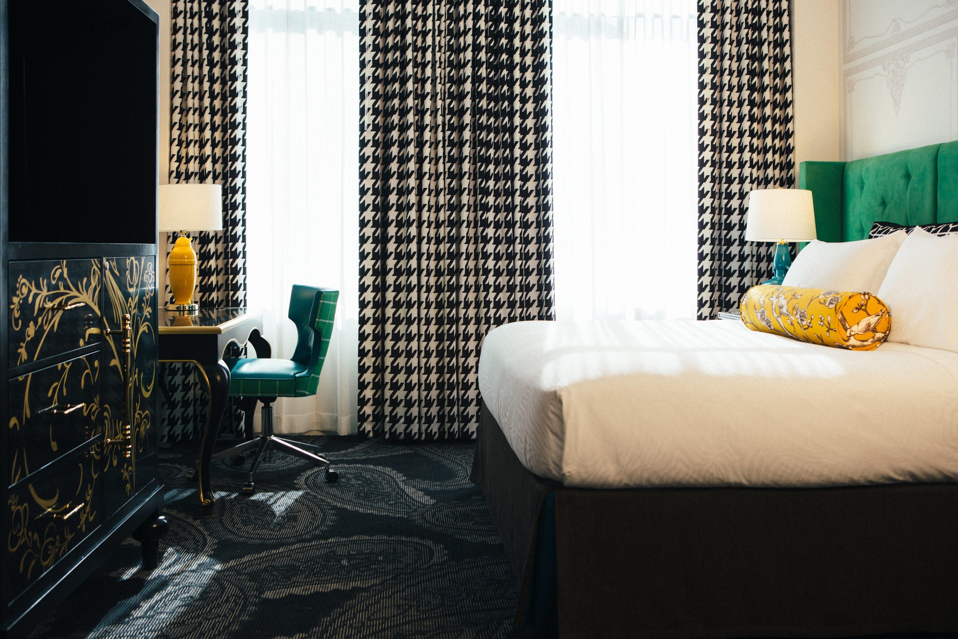 A hotel room with a large bed, a green upholstered chair, a black and gold dresser, and patterned curtains.