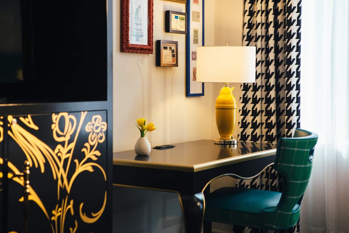 A hotel room with a desk, a lamp, a chair, and a decorative panel.