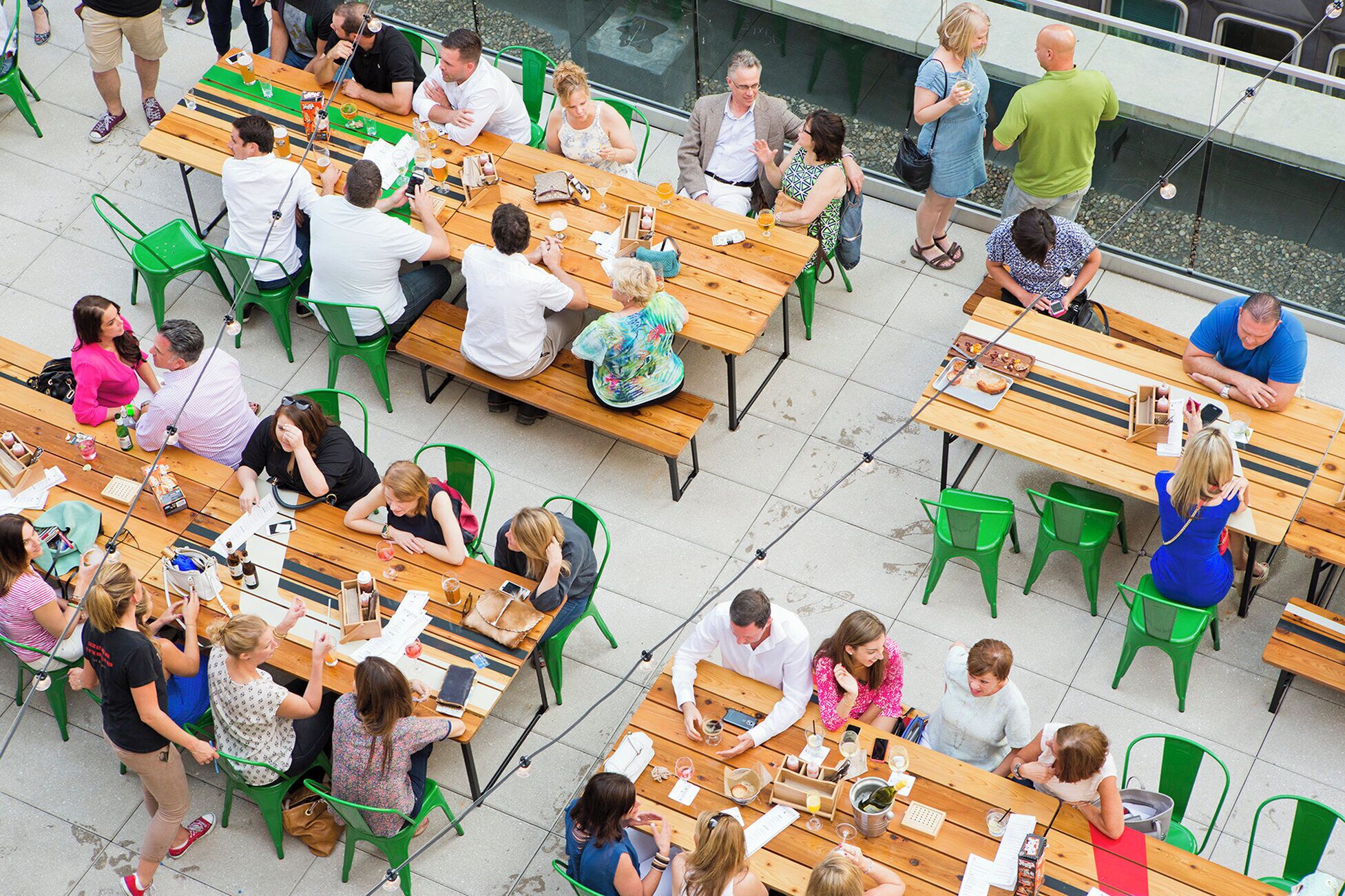 An outdoor dining area with multiple wooden tables and green chairs, surrounded by a glass railing.