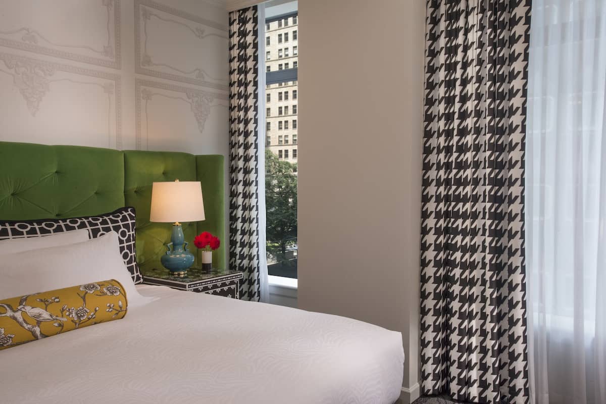 A hotel room with a green upholstered headboard, a white bedspread, a decorative lamp, and a view of a building through sheer curtains.
