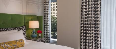 A hotel room with a green upholstered headboard, a white bedspread, a decorative lamp, and a view of a building through sheer curtains.
