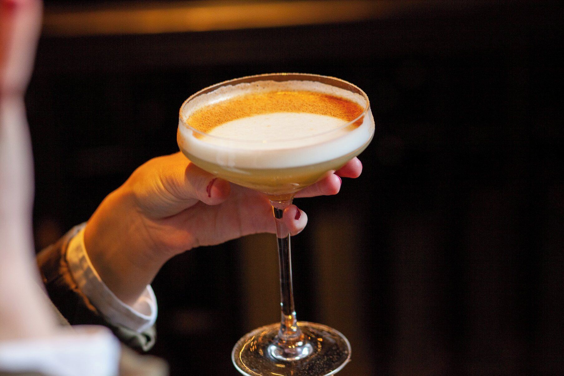 A hand holding a cocktail glass with a creamy top layer, likely a frothy drink.