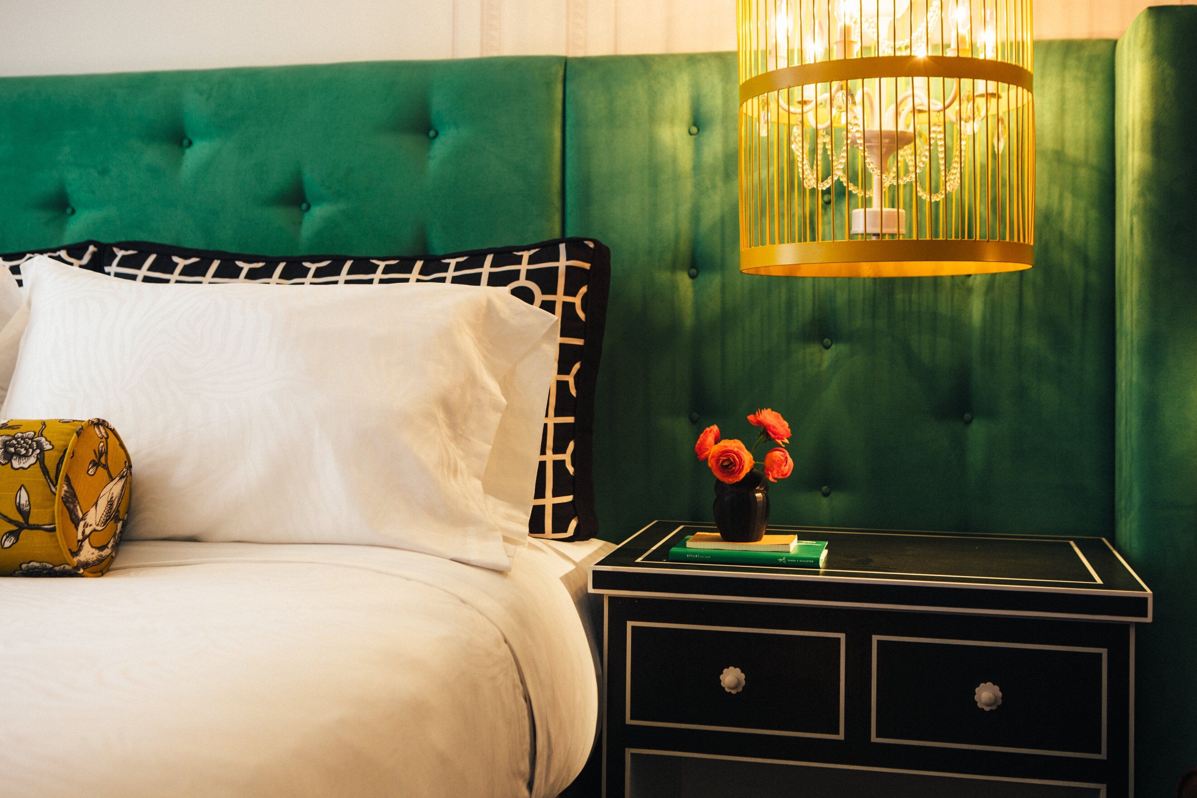 Una cama bien hecha con cabecera tapizada en verde, una mesita de noche con un florero y una lámpara colgante.