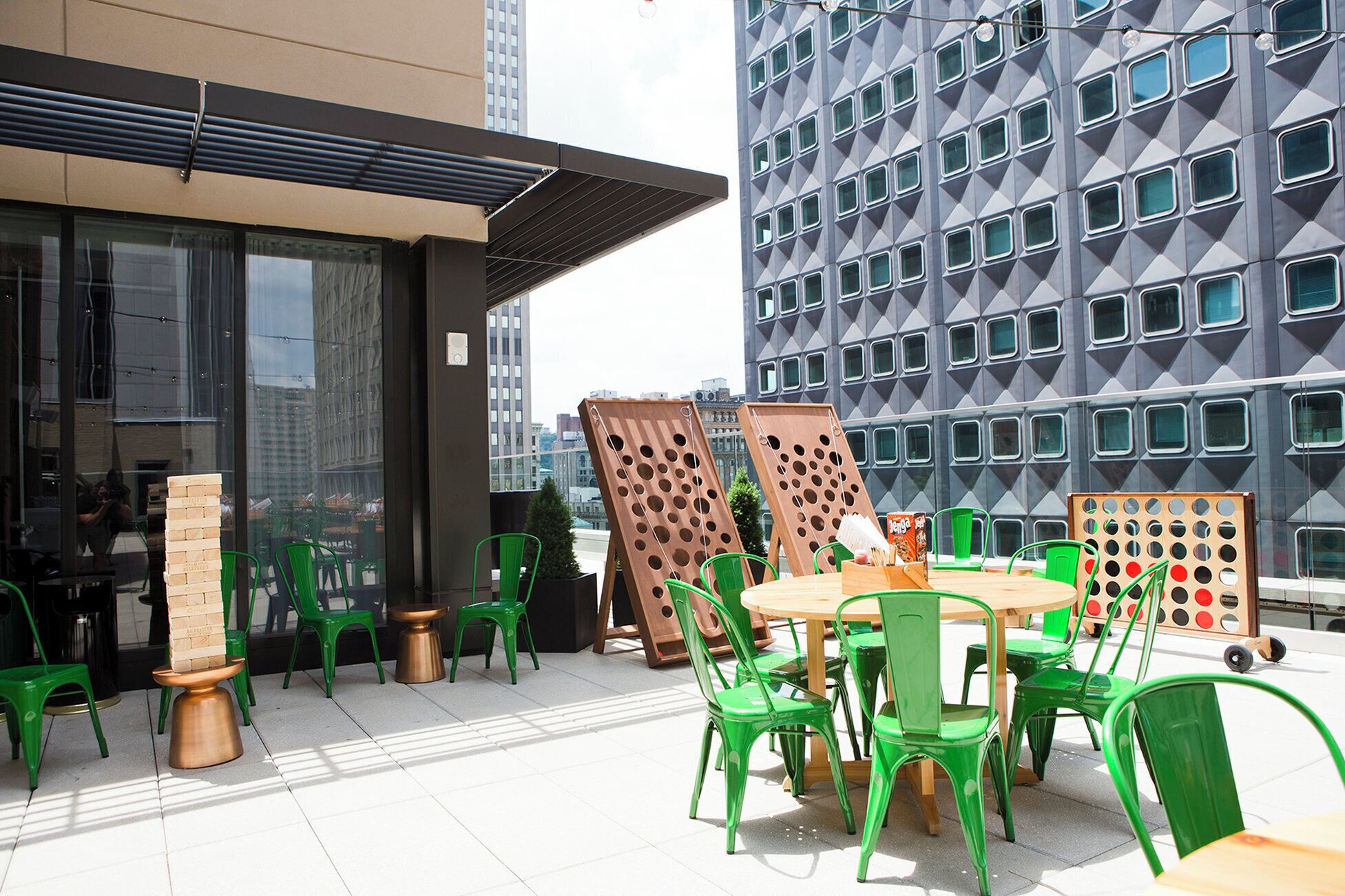 An outdoor seating area with green chairs and a wooden table, overlooking a cityscape with modern buildings.