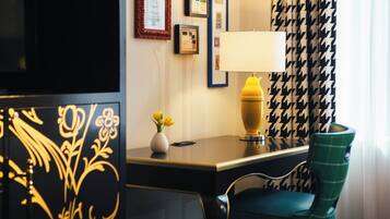 A hotel room with a desk, a lamp, a chair, a vase with flowers, and a patterned curtain.