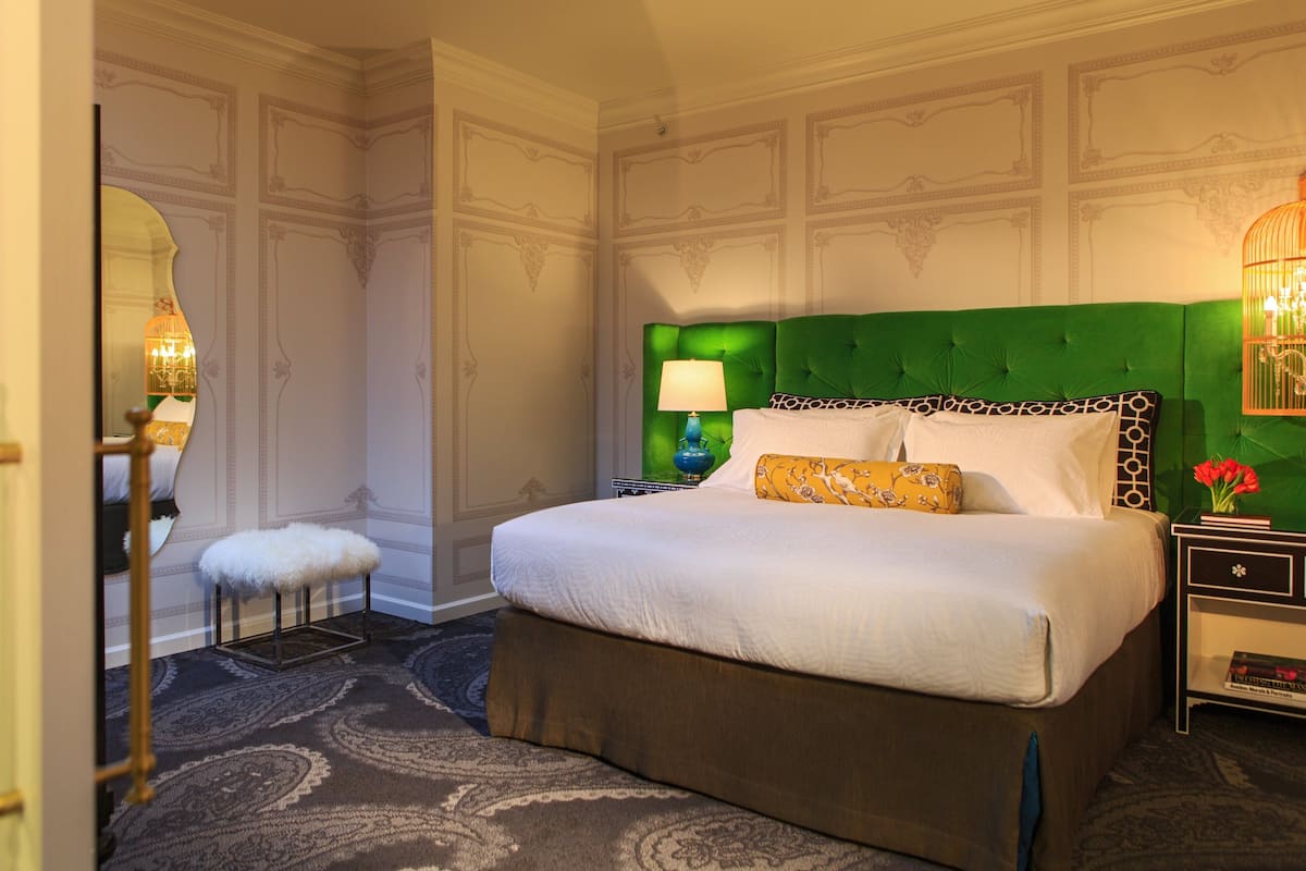 A hotel room with a large bed, a green upholstered headboard, a nightstand with a lamp, a small stool, and a mirror.