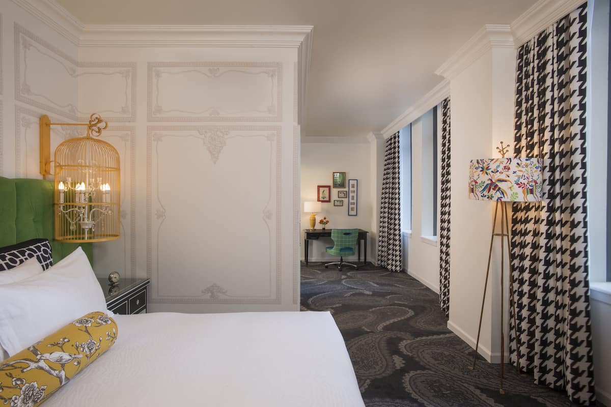 A hotel room with a bed, a green chair, a desk with a lamp, and a houndstooth curtain.