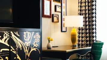 Habitación de hotel con un mueble negro decorado con motivos florales dorados, un escritorio con lámpara, una silla verde y cortinas estampadas.