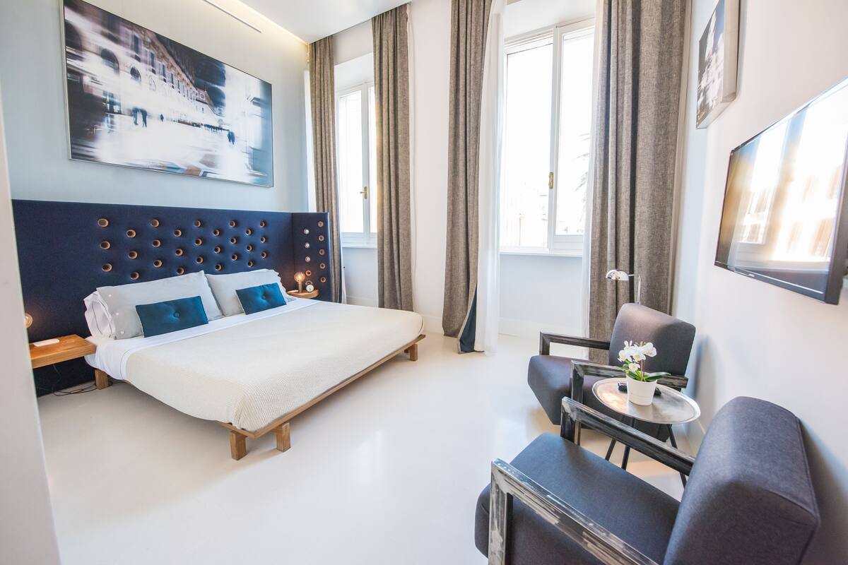 A modern hotel room with a large bed, a seating area with a small table, and a large window with curtains.