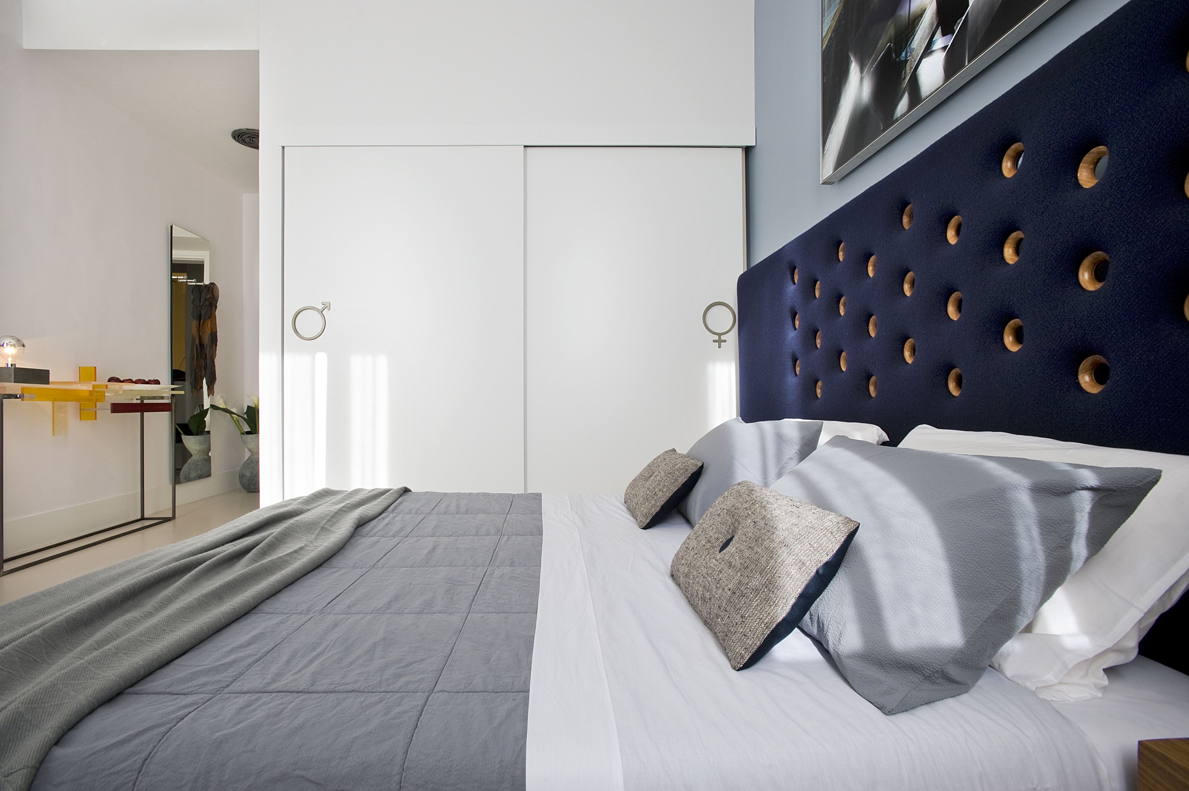 A modern bedroom with a grey bed, white bedding, and a blue headboard with circular cutouts.