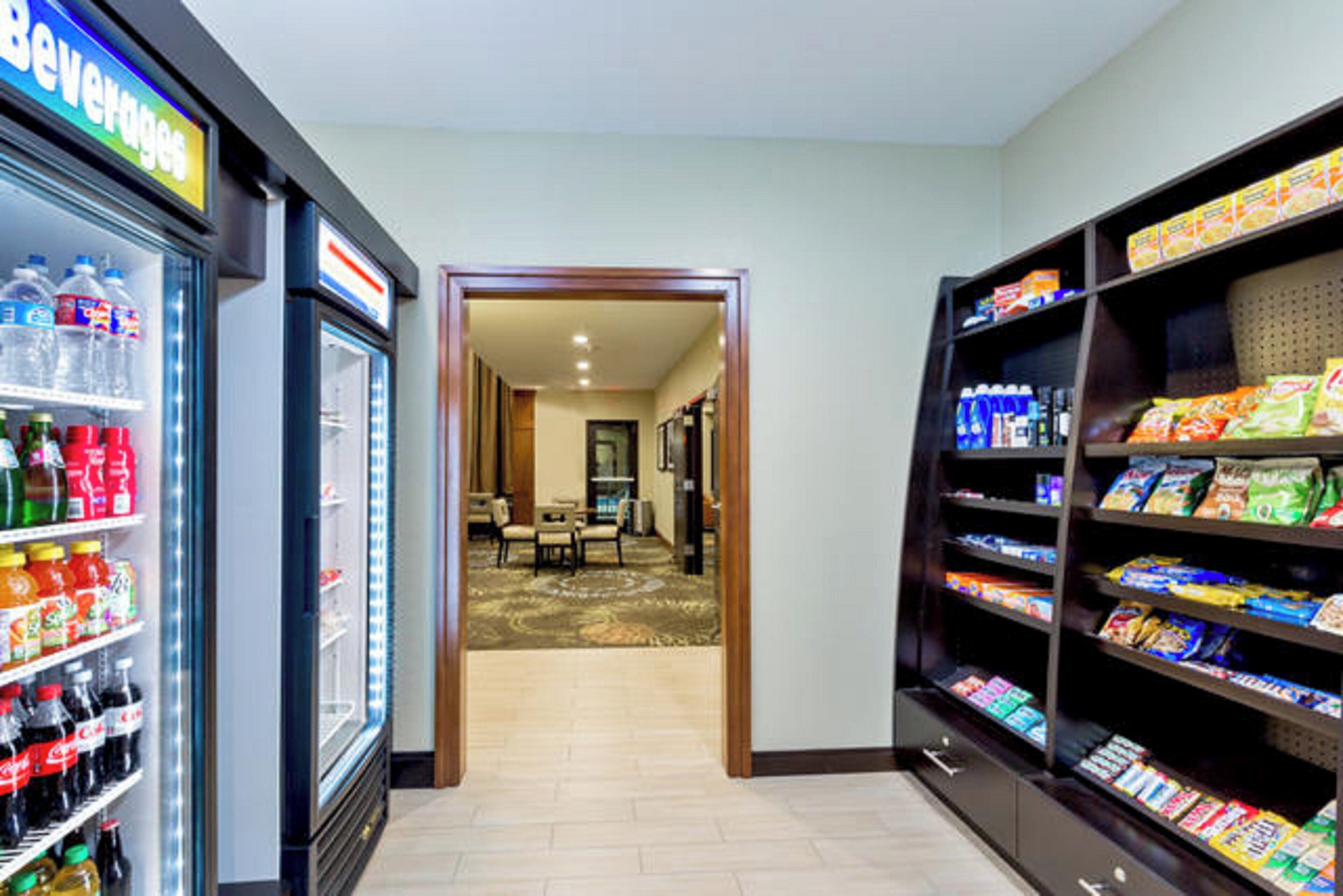 A hotel lobby with a beverage refrigerator, snack shelves, and a carpeted area leading to a room.