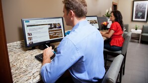 A man writing at a desk with a computer displaying a webpage. Another person is seated at a table in the background. - Staybridge Suites College Station by IHG (College Station)