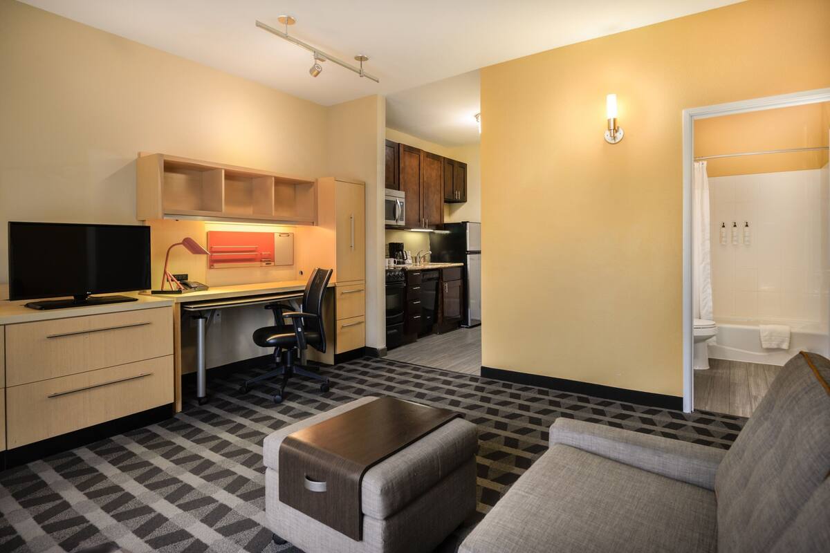 Ein kompaktes Hotelzimmer mit Küchenzeile, einer Couch, einem Schreibtisch mit Stuhl und einem Fernseher.