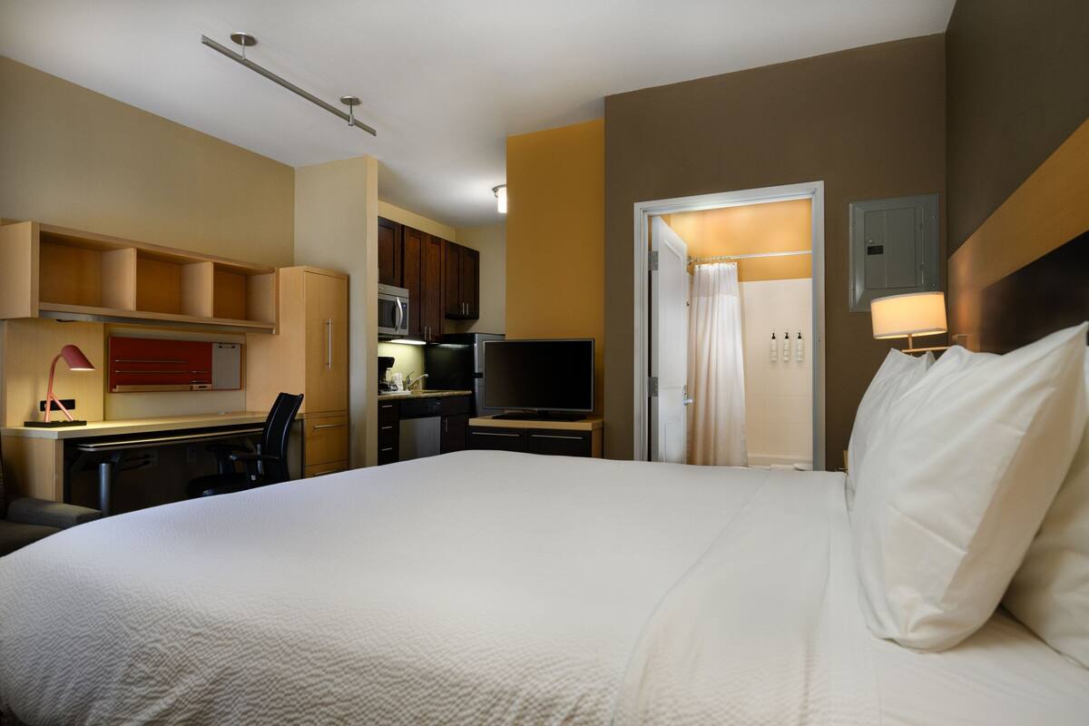 Ein Hotelzimmer mit einem großen Bett, einem Schreibtisch mit Computer, einem Fernseher und einer Küchenzeile.