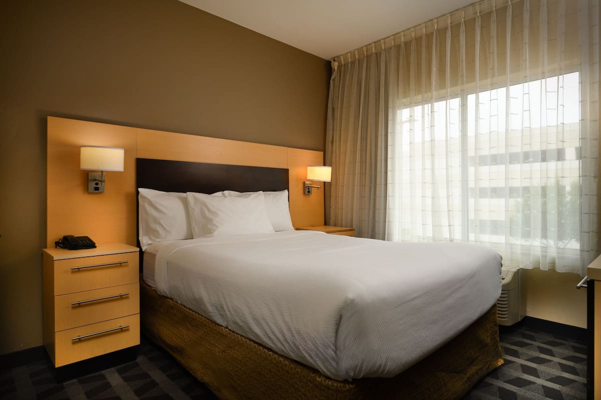 Ein Hotelzimmer mit einem großen Bett, zwei Nachttischen mit Lampen, einem Holzbeistelltisch und einem Fenster mit leichtem Vorhang.