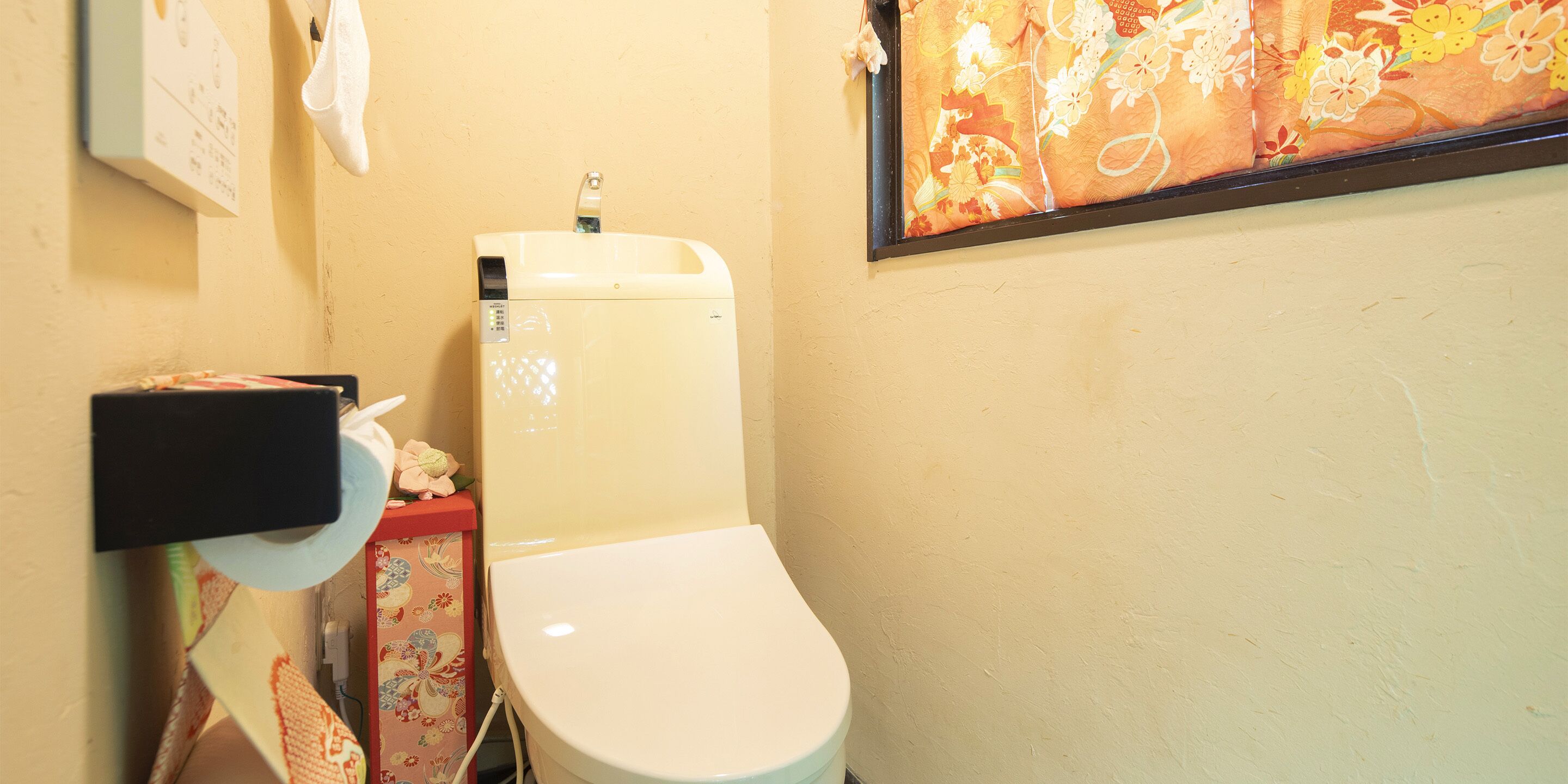 Sanitário com assento e caixa de descarga brancos, dispensador de papel e uma pequena lixeira.