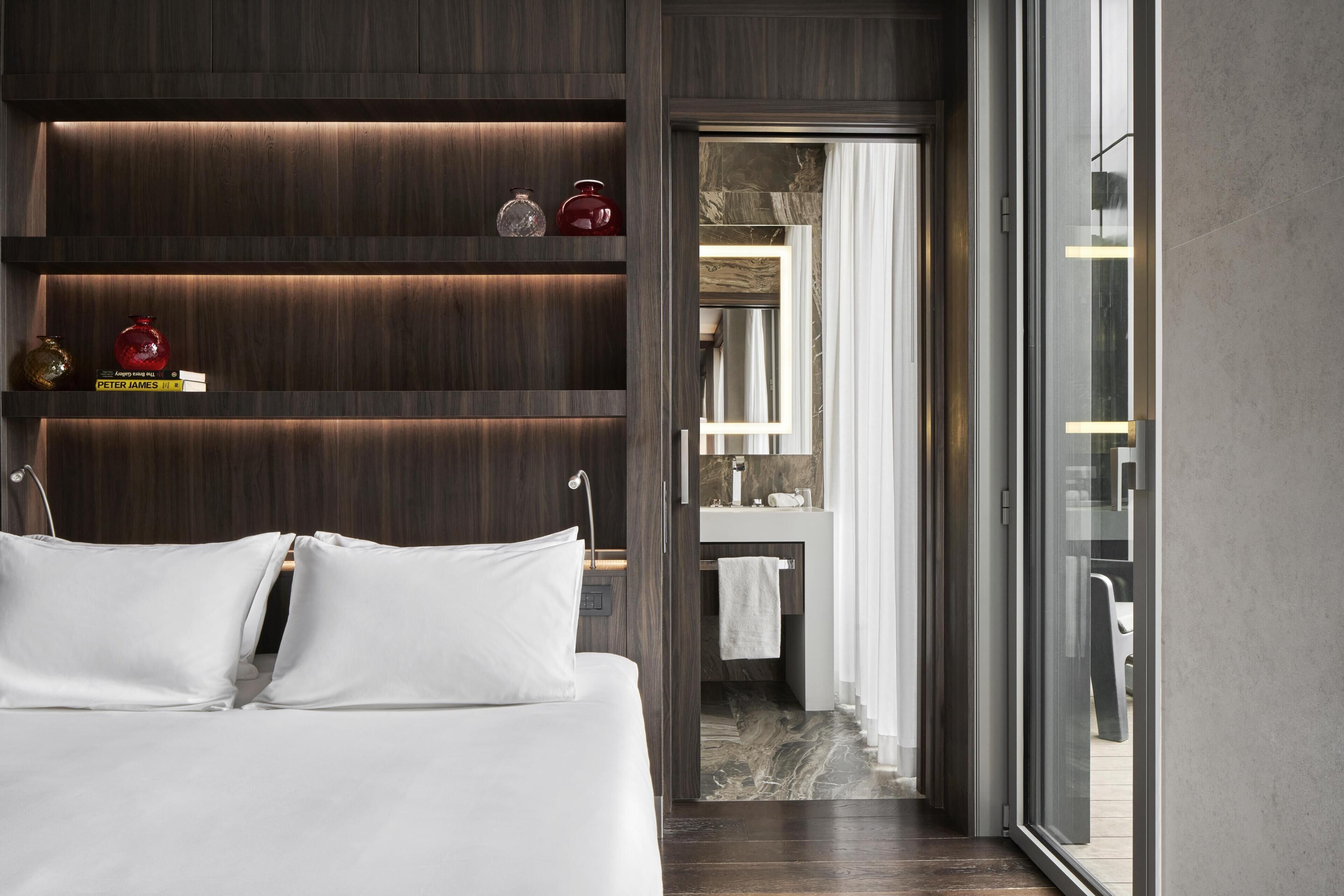 A modern hotel room with a large bed, a wooden shelf with decorative items, a bathroom with a mirror and sink, and a view through a glass door.