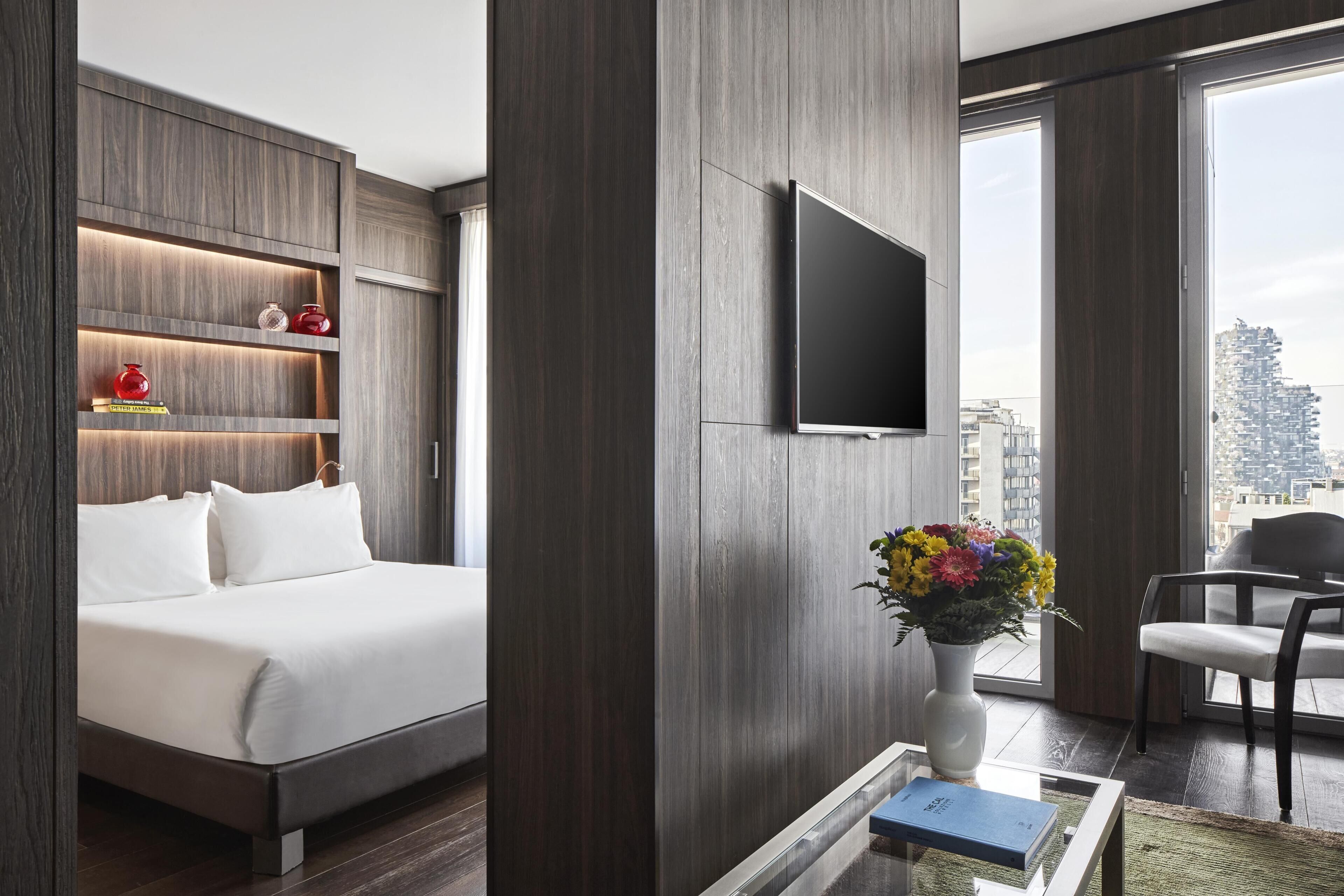 Quarto de hotel moderno com cama, televisão de tela plana, um vaso com flores e vista para a cidade.