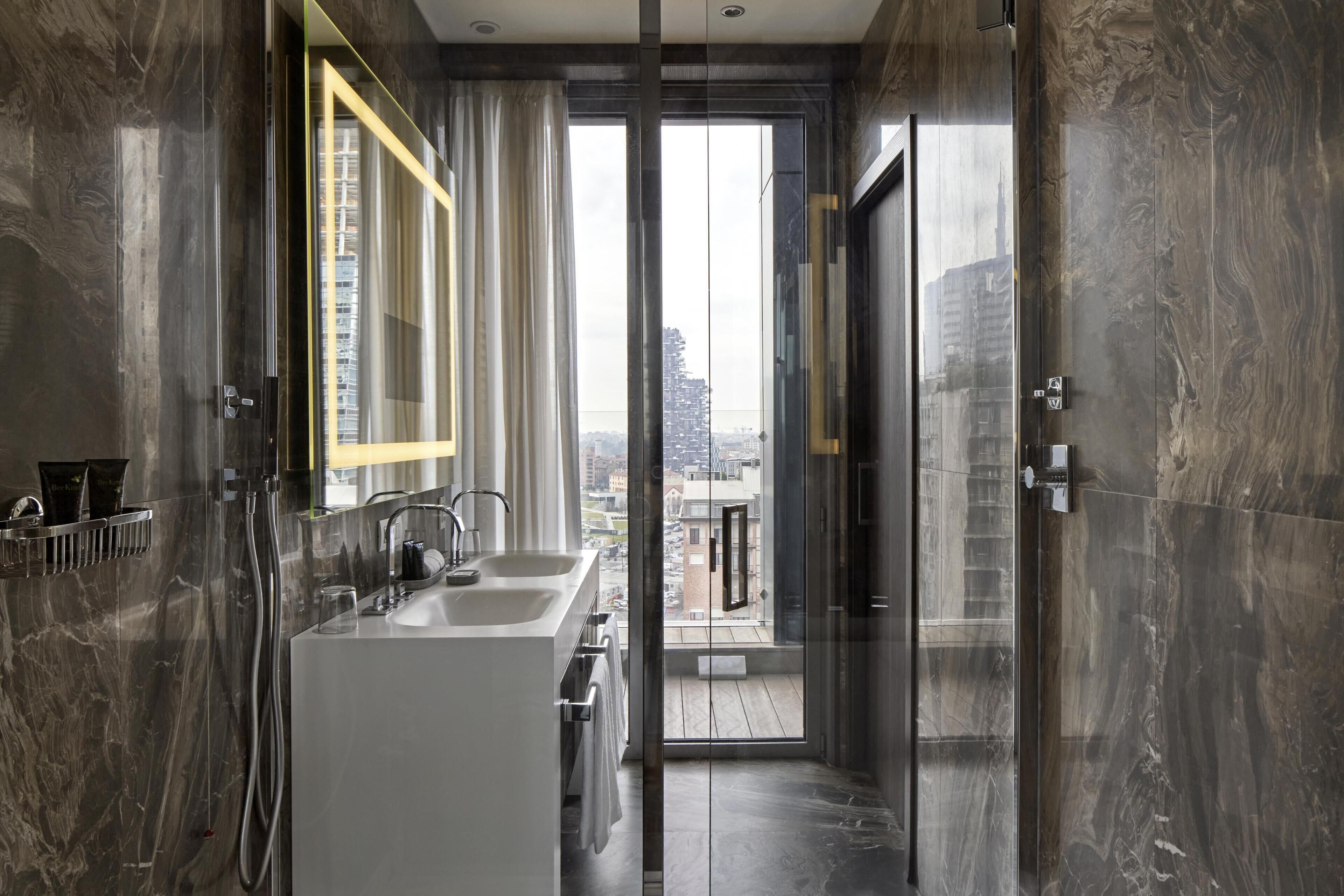 A modern bathroom with a dual sink, a glass shower enclosure, and a view of the cityscape through a large window.