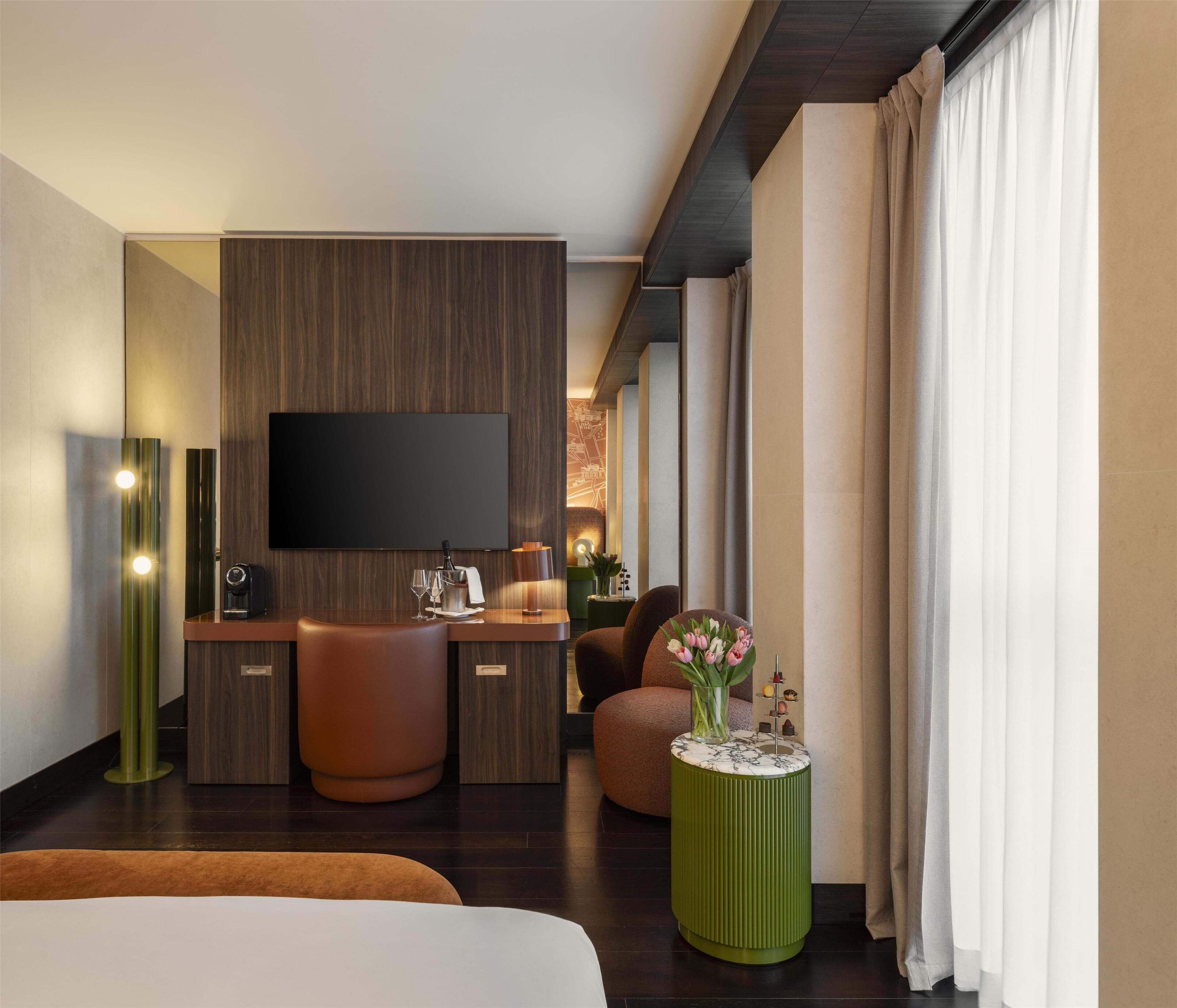 Een moderne hotelkamer met een flatscreen televisie, een houten bureau, een comfortabele zithoek en een groot raam met gordijnen.