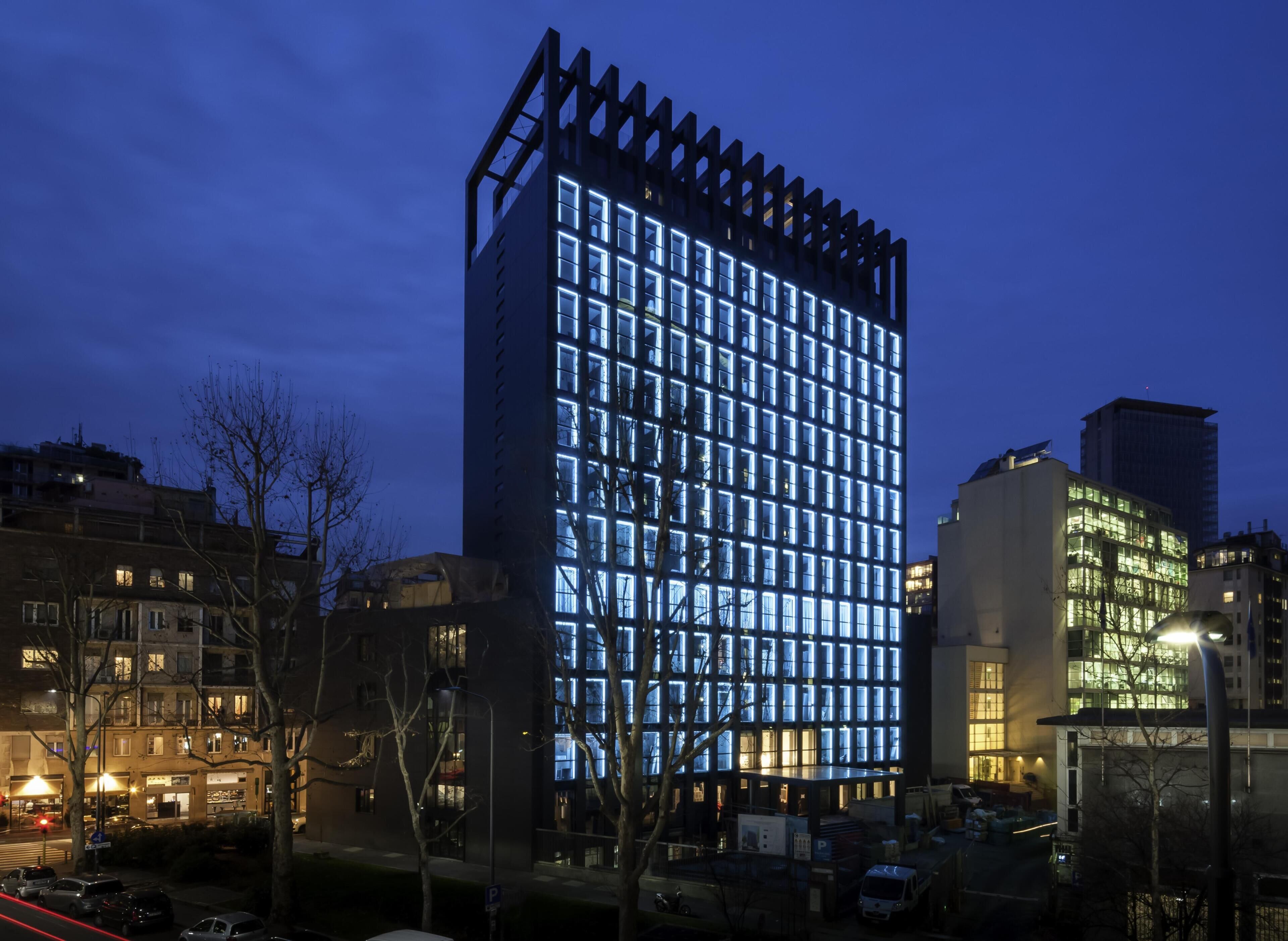 Un moderno edificio de gran altura con una cuadrícula de ventanas iluminadas, rodeado de otros edificios y automóviles estacionados.