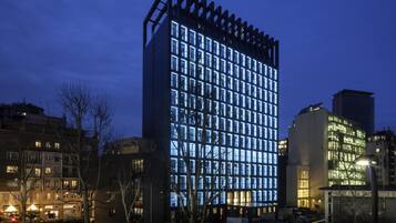 Un moderno edificio de gran altura con una cuadrícula de ventanas iluminadas, rodeado de otros edificios y automóviles estacionados.