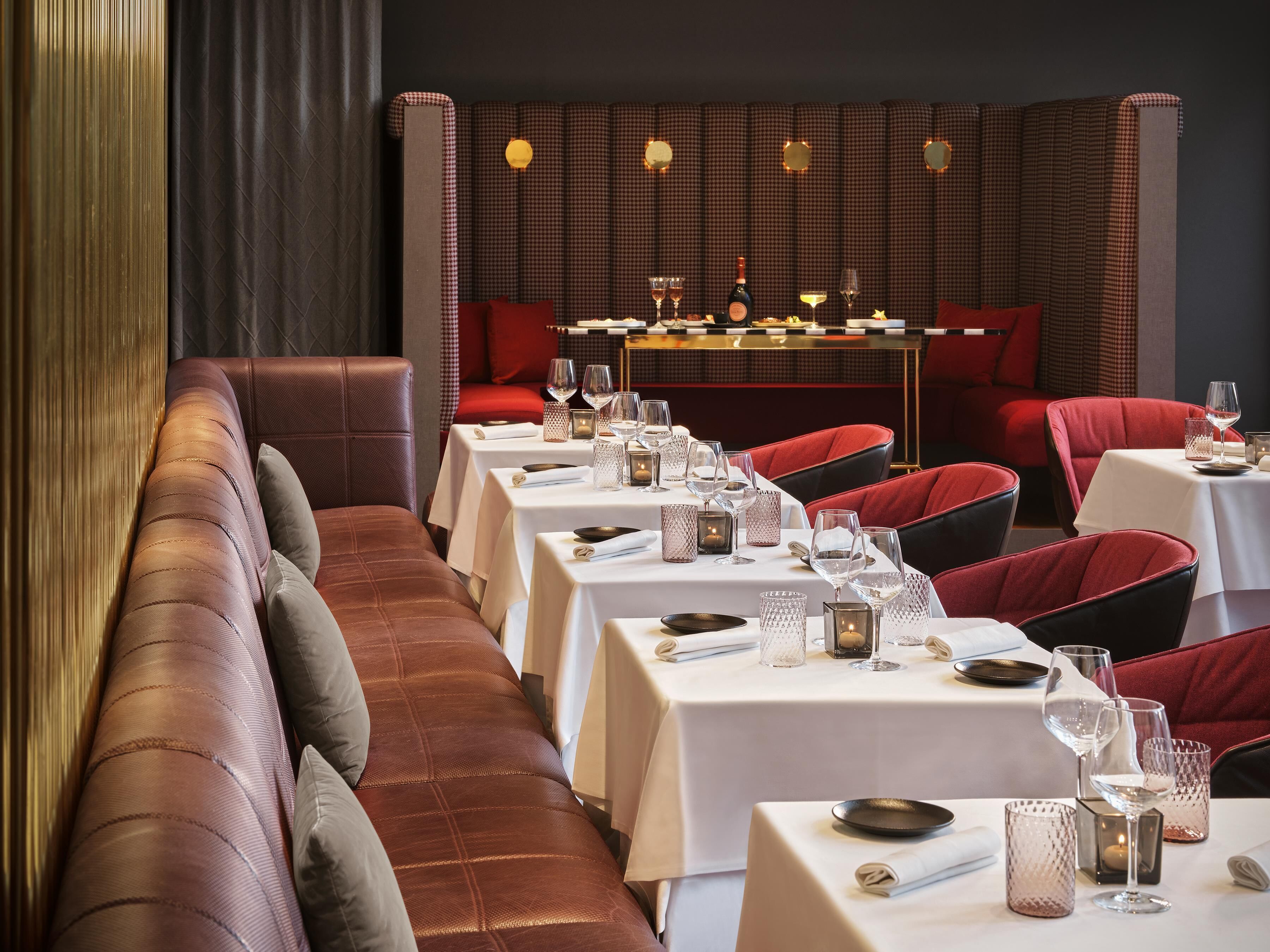 A restaurant with a long bench seating area, tables with white tablecloths, and red upholstered chairs.