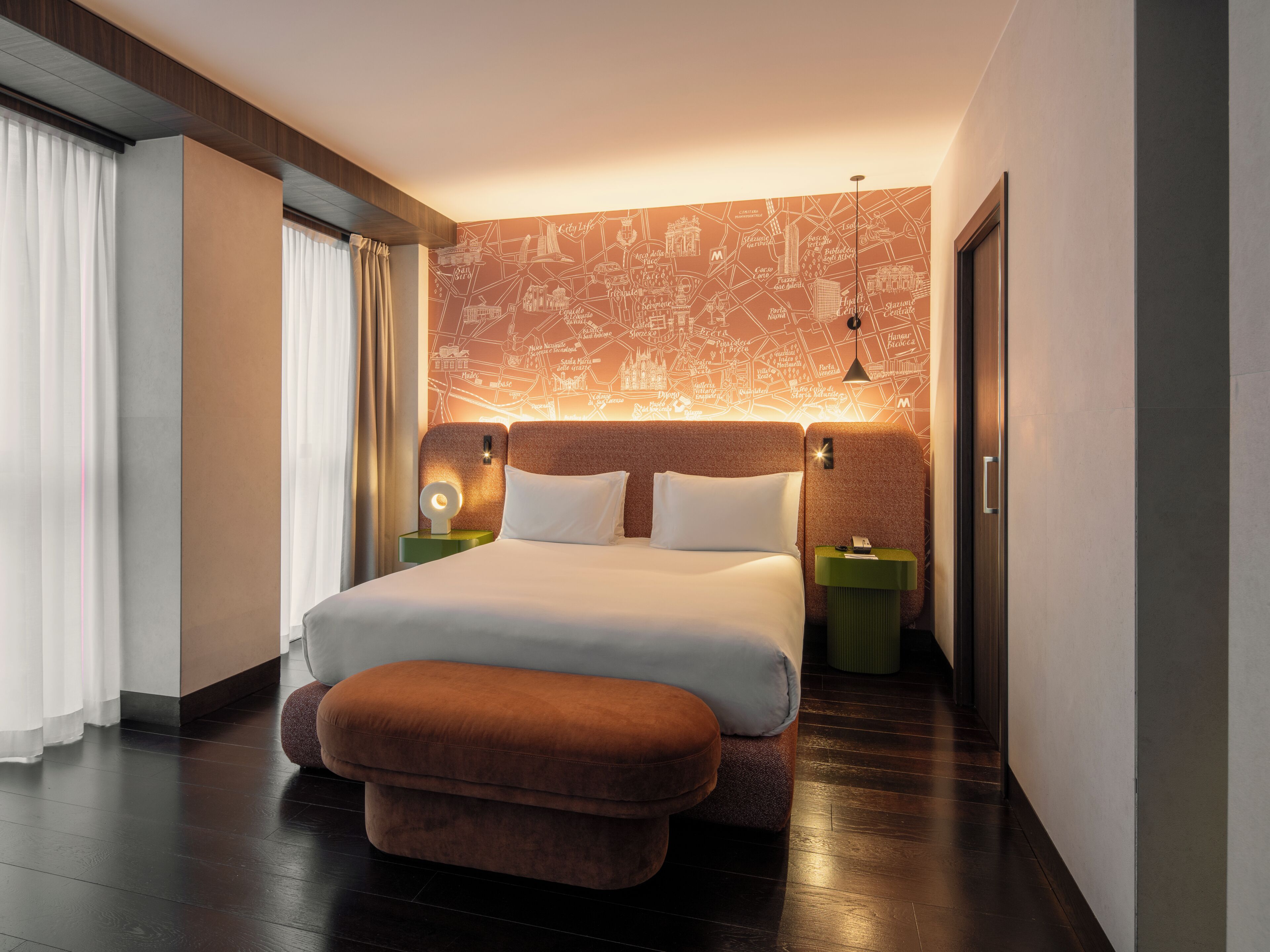 A modern hotel room with a large bed, a nightstand, a green side table, a brown ottoman, and a wall with a cityscape map.