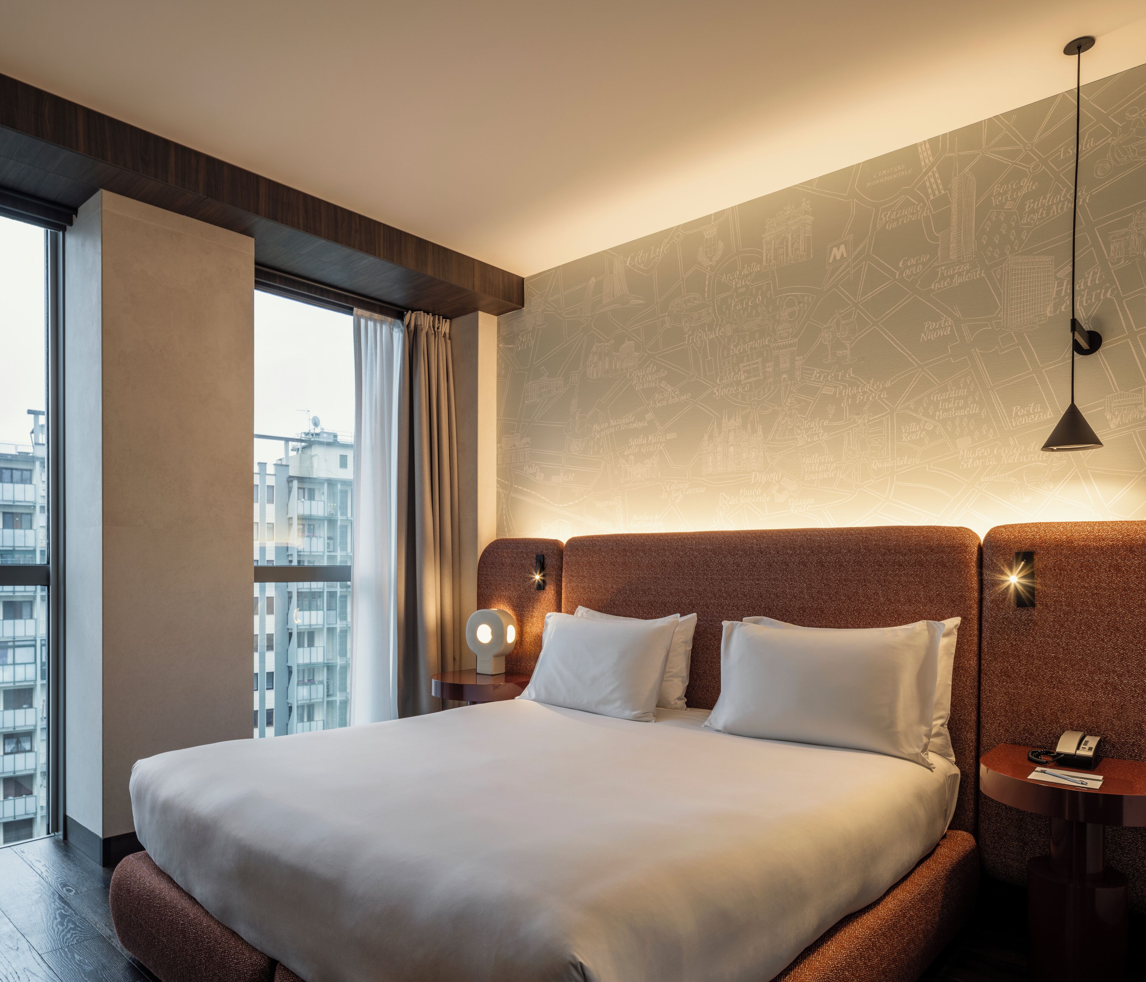Habitación de hotel con una cama grande, una mesita de noche, una lámpara y vistas a la ciudad a través de una ventana.