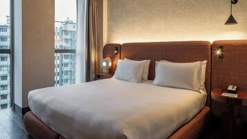 Habitación de hotel con una cama grande, una mesita de noche, una lámpara y vistas a la ciudad a través de una ventana.