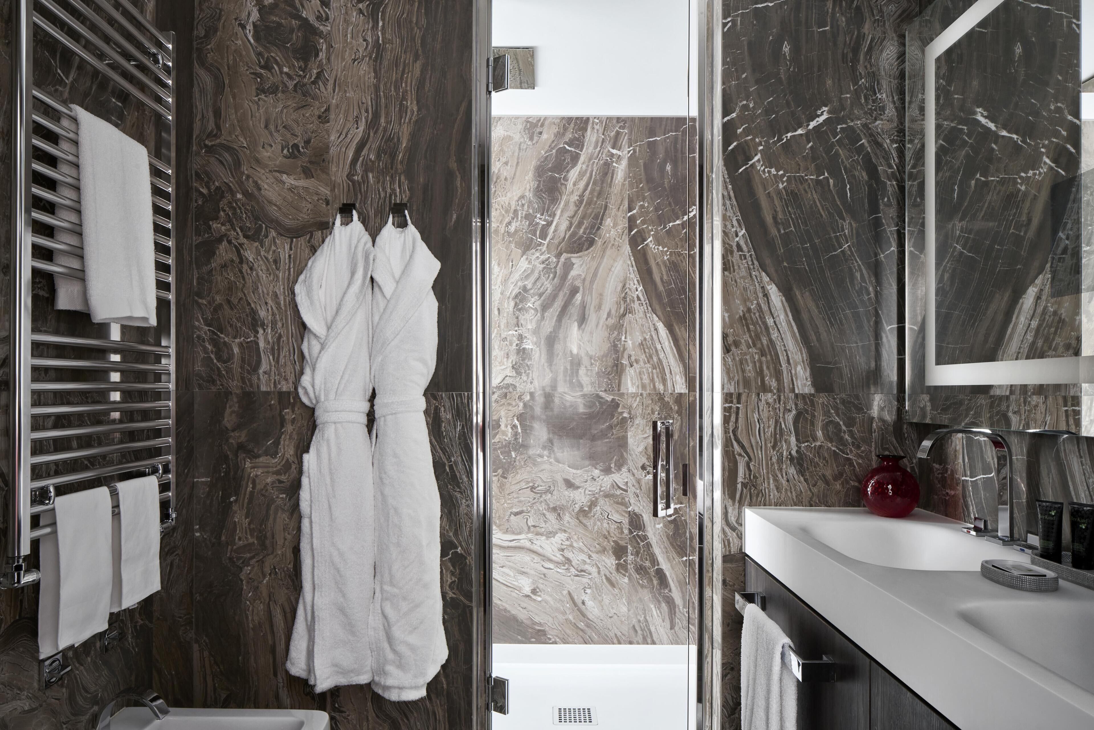 A modern bathroom with a marble wall, a white bathtub, a glass shower enclosure, and a white robe hanging on a rack.