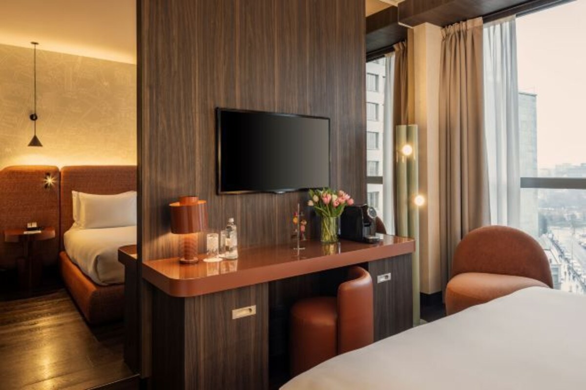 Habitación de hotel con cama, escritorio con televisor de pantalla plana, jarra con flores, cafetera y vista a la ciudad.