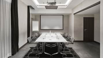 Uma sala de conferências com uma mesa grande, cadeiras, uma tela de projetor e um quadro branco.