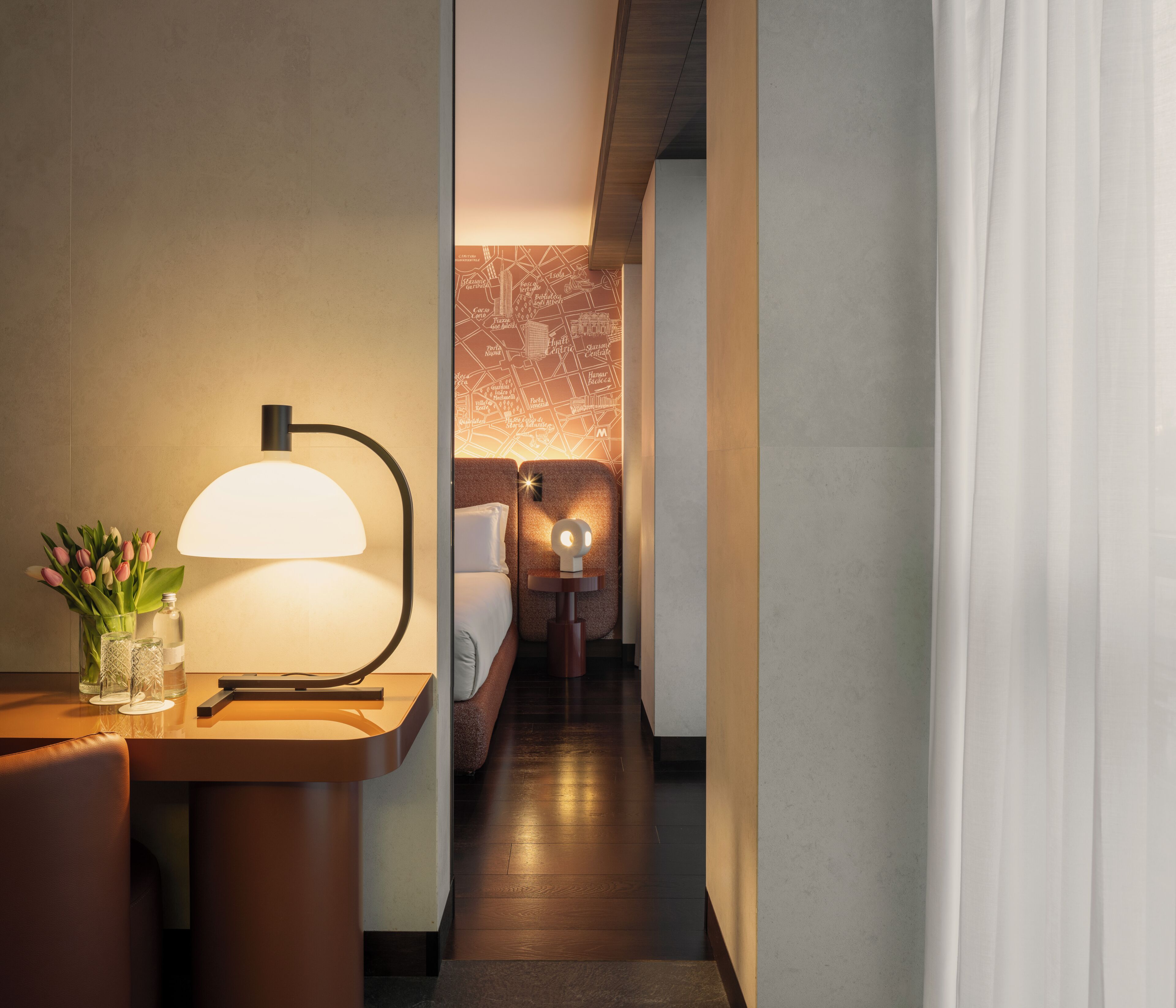 A hotel hallway with a lamp, a table with a vase of flowers, and a bed in the background.