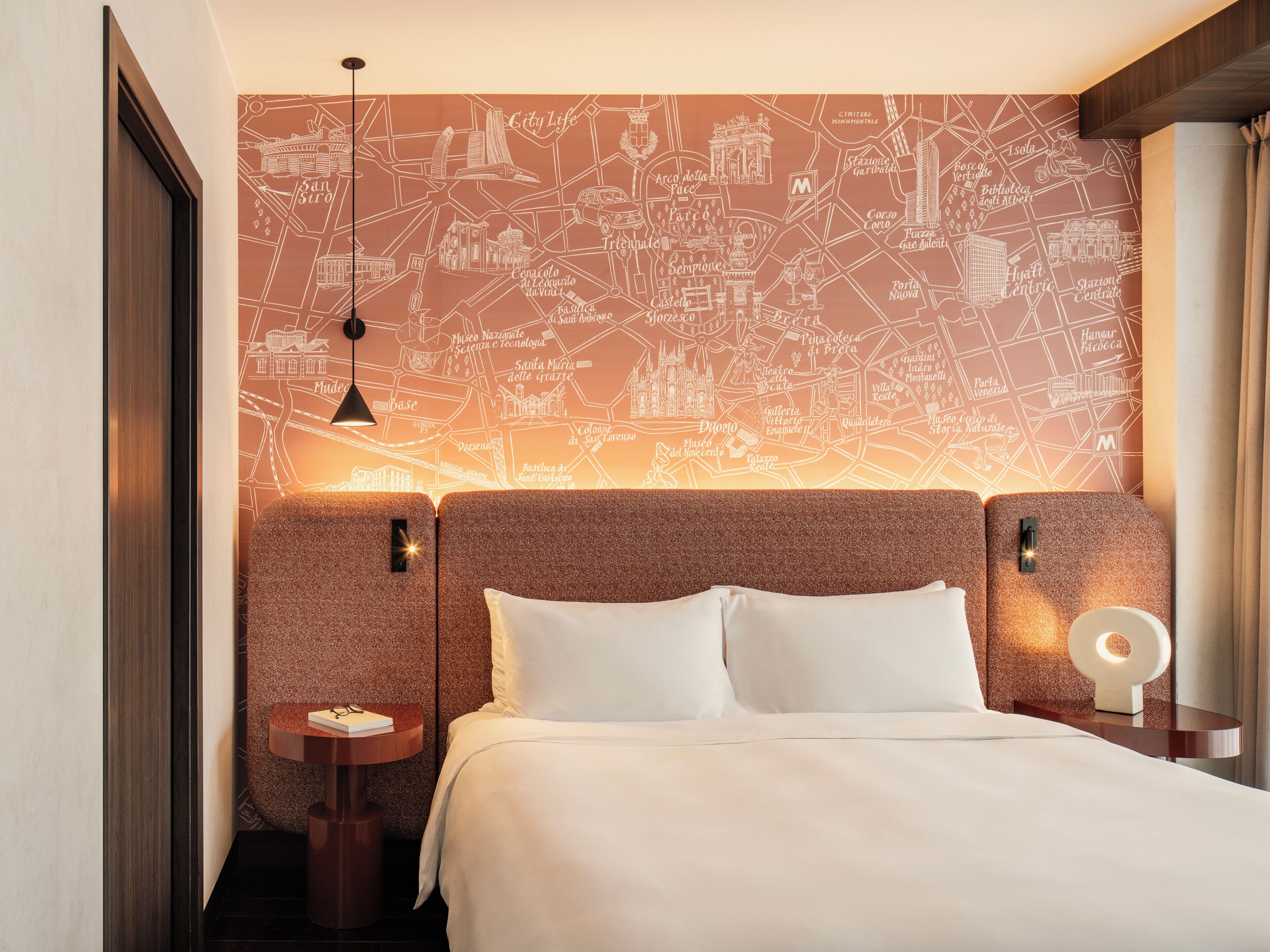 Hotelkamer met stadsgezicht aan de muur, een bed met witte lakens, twee nachtkastjes en een hanglamp.